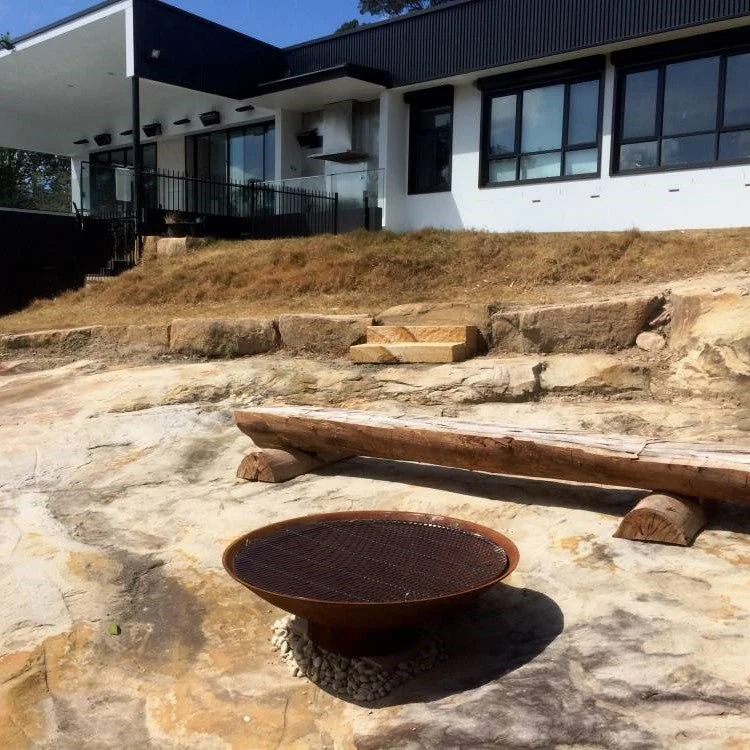A large round cast iron fire pit, with stainless steel cooking grill on a large, cemented area, a large wooden sleeper for seating and a house in the backdrop.