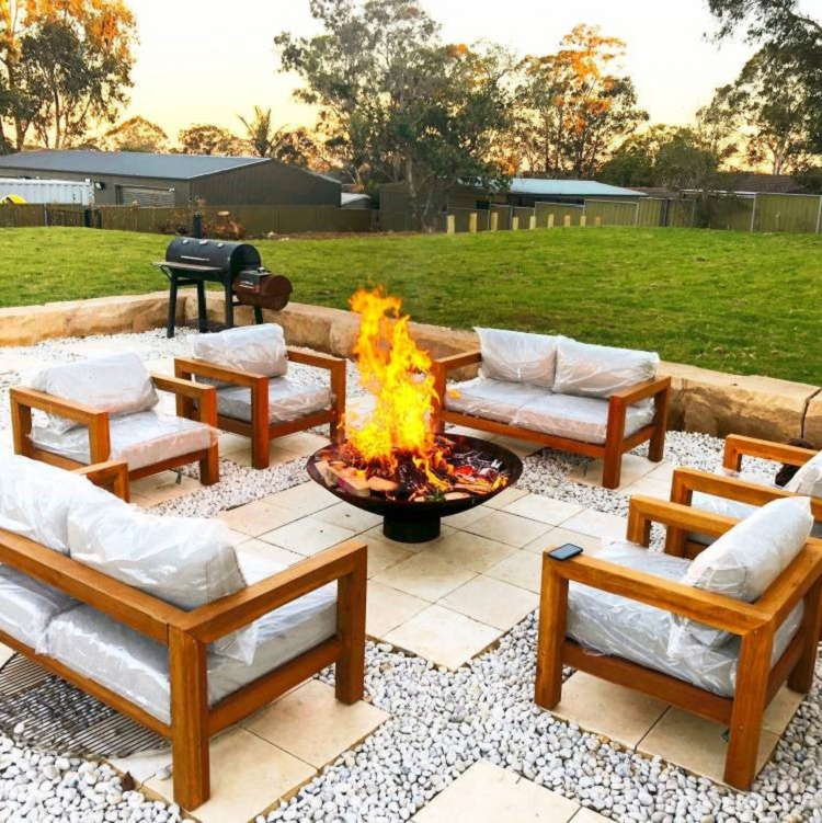 A large round cast iron fire pit, with fire burning in a backyard setting with pavers, pebbles and outdoor seating cemented area with a large lawn area, fencing and trees in the backdrop.