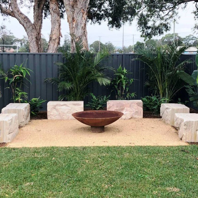 A round, rust-colored cast iron fire pit, placed on a bed of sand, in a garden setting. There are six large stone slabs for sitting around the fire pit.