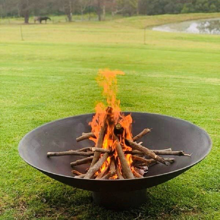 A round, rust-colored cast iron fire pit, with a small fire burning placed on a large lawn area with a lake, trees and a couple of horses nearby in the background.