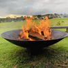 A round, rust-colored cast iron fire pit, with fire burning placed on a large lawn area with a house, trees and a couple of sheds in the nearby background.