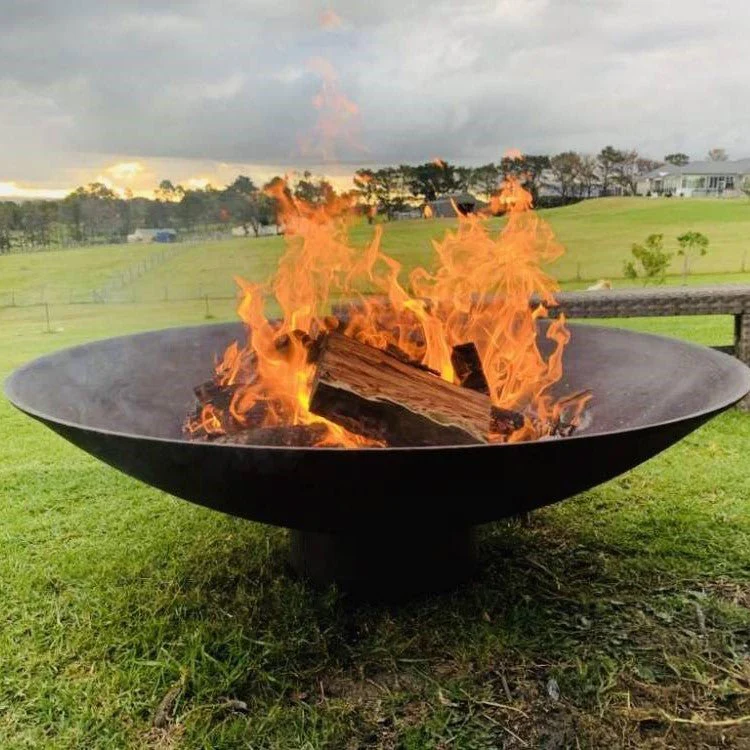 A round, rust-colored cast iron fire pit, with fire burning placed on a large lawn area with a house, trees and a couple of sheds in the nearby background.