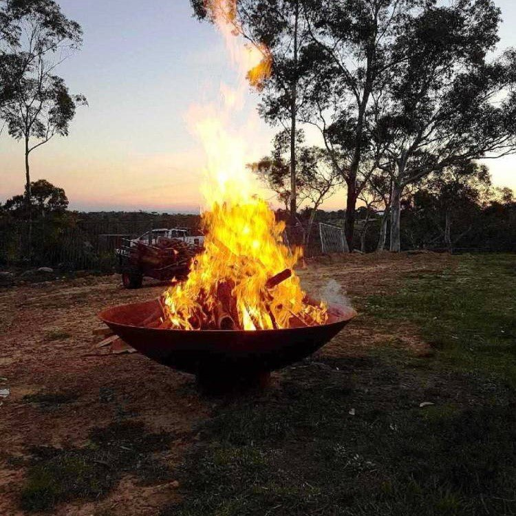 Large round, rust-colored cast iron fire pit, with fire burning placed on a large sandy lawn area with a trailer, trees and a ute close by.