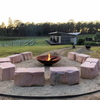 A large round, rust-colored cast iron fire pit, with fire burning placed on a gravel area with ten large stone slabs for sitting around the fire. There is a house, trees and vineyard in the nearby setting.