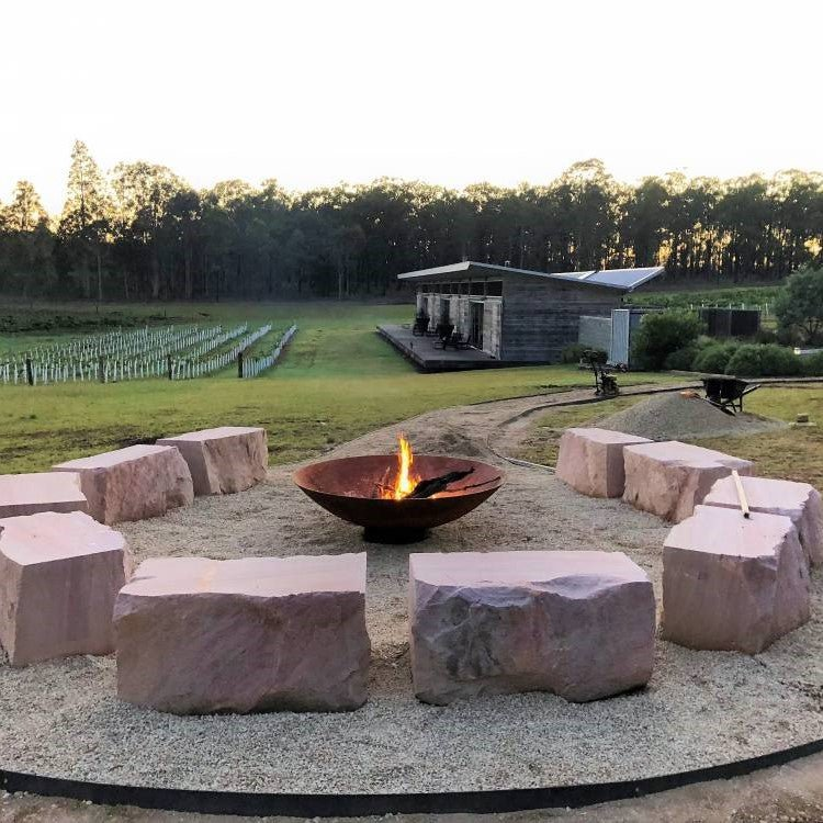 A large round, rust-colored cast iron fire pit, with fire burning placed on a gravel area with ten large stone slabs for sitting around the fire. There is a house, trees and vineyard in the nearby setting.