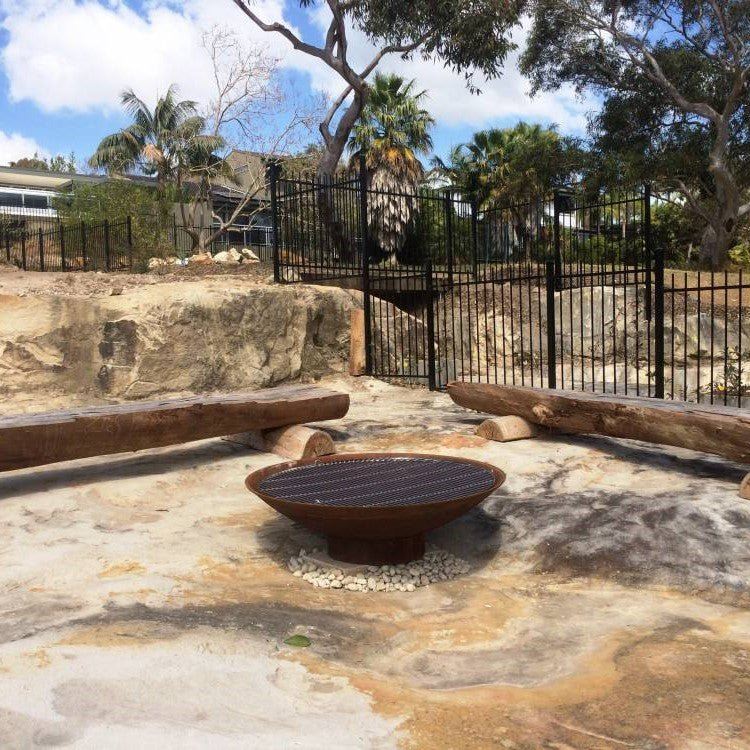 A round, rust-colored cast iron fire pit, with stainless steel grill placed on a large, cemented garden area with a house, trees and fences in the background.