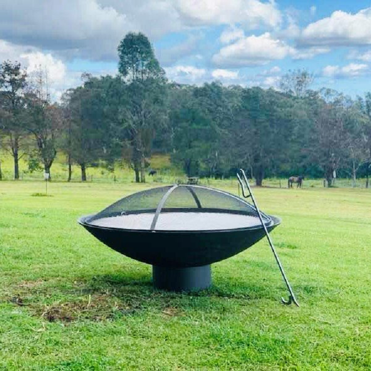 A large round cast iron fire pit, with ember screen on it and a metal poker leaning on it placed on a large, lawn area with a couple of horses and trees in the backdrop.