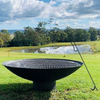 A large round cast iron fire pit, with metal grill on top of it and a metal poker leaning on it placed on a large, lawn area with a lake and trees in the backdrop.