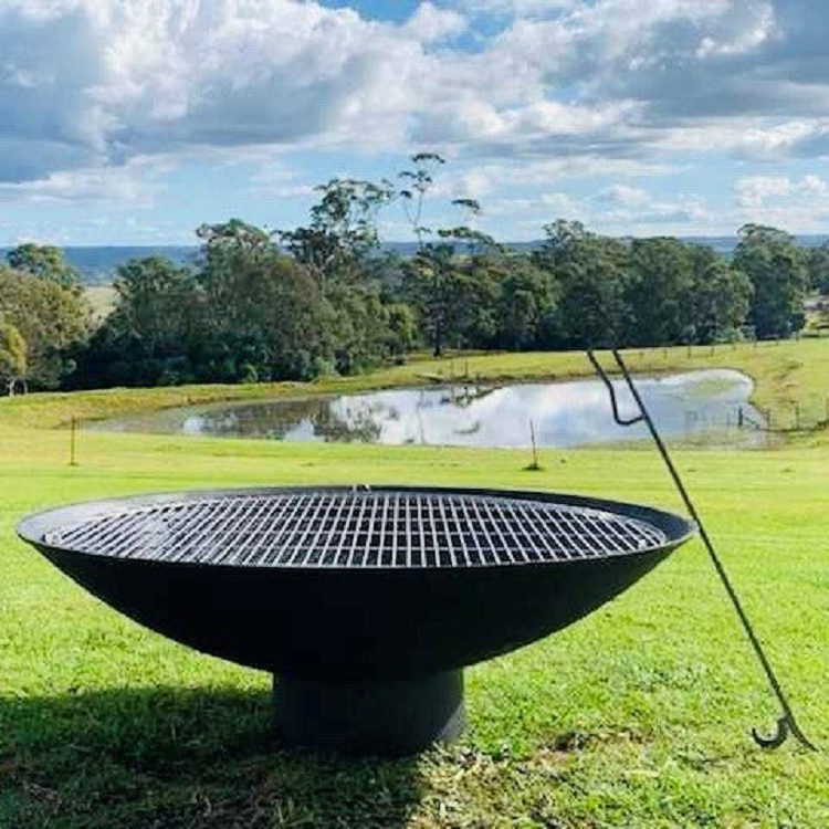A large round cast iron fire pit, with metal grill on top of it and a metal poker leaning on it placed on a large, lawn area with a lake and trees in the backdrop.