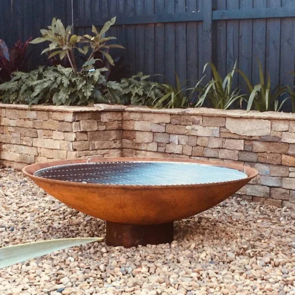 A round, rust-colored cast iron fire pit, placed on a bed of pebbles, set against a garden backdrop. The fire pit has a smooth, solid appearance with a slightly elevated rim.