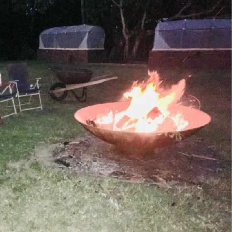 Large size Cauldron Fire Pit at a camp site with fire burning, a couple of camp chairs and a wheelbarrow sitting next to the burning fire.