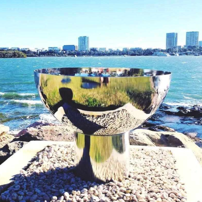 The Goblet Stainless Steel Fire Pit beside a body of water with trees and buildings in background