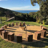 The Goblet Stainless Steel Fire Pit in the middle of log seats with trees and distant ocean view in background