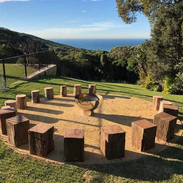 The Goblet Stainless Steel Fire Pit in the middle of log seats with trees and distant ocean view in background