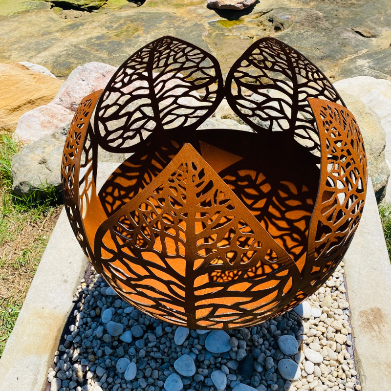 Top view of the Leaf Fire Pit showing the intricate vein design, in natural rust colour. Sitting on a pebble surround and a rocky ground.