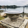 Teppanyaki Stainless Steel Fire Pit complete with BBQ grill, brackets, spit poles and rotisserrie besides the water on a gravel and rocky surrounding.
