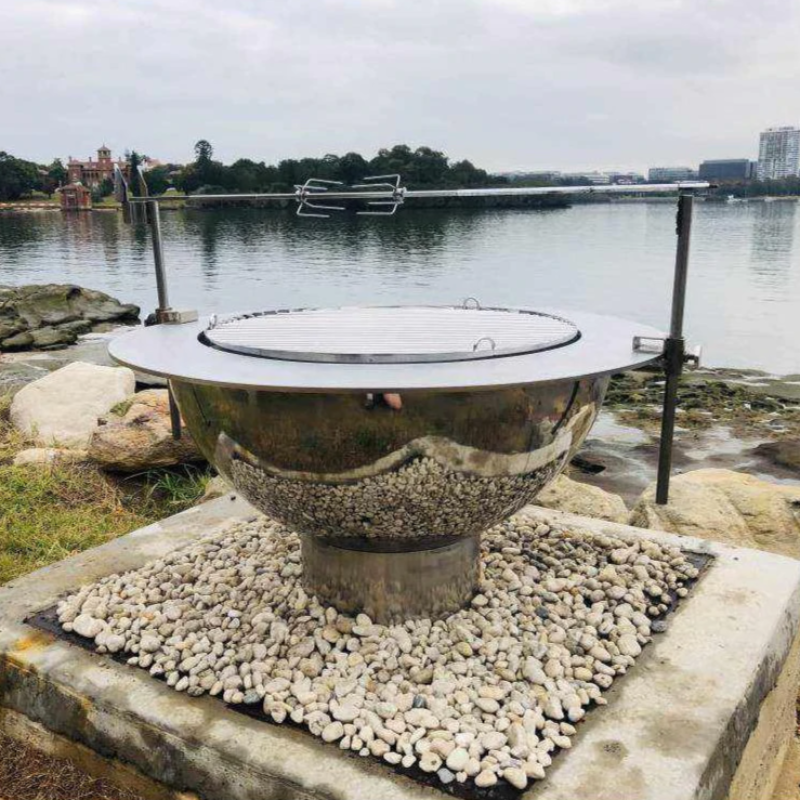 Teppanyaki Stainless Steel Fire Pit complete with BBQ grill, brackets, spit poles and rotisserrie besides the water on a gravel and rocky surrounding.