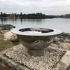 The Teppanyaki Stainless Steel Fire Pit besides a body of water on a gravel and rocky surround with trees and buildings in background.