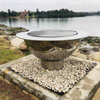 The Teppanyaki Stainless Steel Fire Pit and grill besides a body of water on a gravel and rocky surround with trees and buildings in background.
