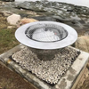 The Teppanyaki Stainless Steel Fire Pit and grill set up on a gravel and rocky surrounding.