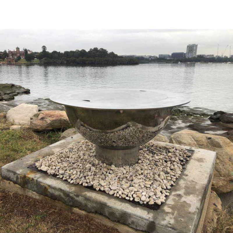 The Teppanyaki Stainless Steel Fire Pit and lid on set up beside a body of water on a gravel and rocky surrounding.