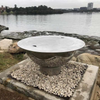 The Teppanyaki Stainless Steel Fire Pit and lid on set up beside a body of water on a gravel and rocky surrounding with trees and buildings in background.
