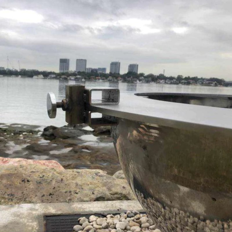 Close up of custom-made brackets on the Teppanyaki Stainless Steel Fire Pit beside the water with trees and buildings in background.