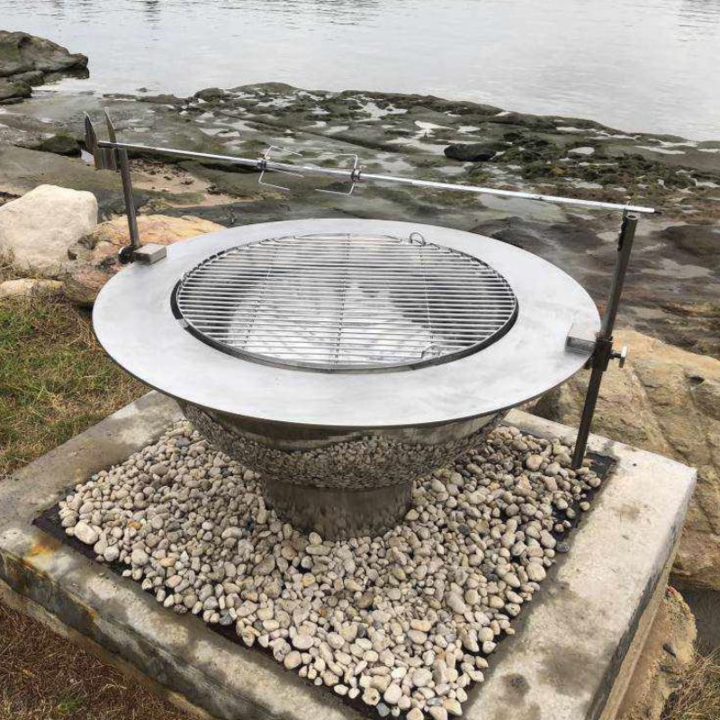 Complete set up of the Teppanyaki Stainless Steel Fire Pit with gravel and rocky surrounding beside water.