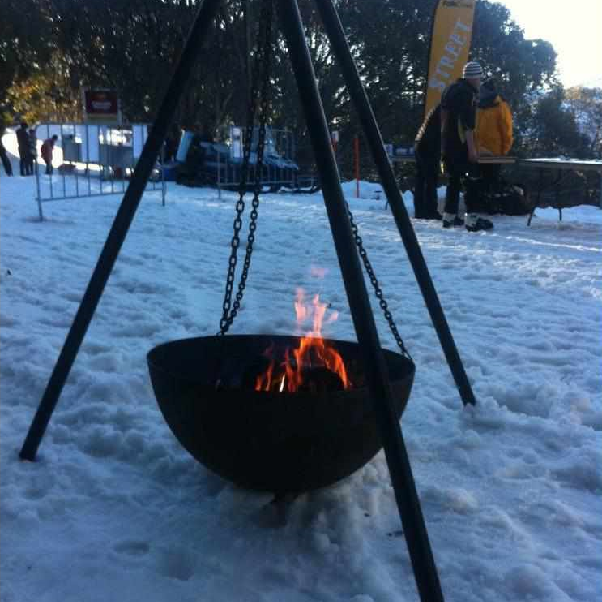 The Tripod Fire Pit in the snow with a burning fire, people and trees in the background.