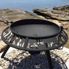 The Anzac patterned Ultimate BBQ Fire Pit with the words 'Lest We Forget' and soldiers cut out, set up on a gravel and rocky surround besides a lake