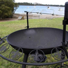 The Ultimate BBQ Fire Pit with a spit rotisserie and the Fleur De Lis pattern, on a lawn area beside a body of water with boats and trees in the background.