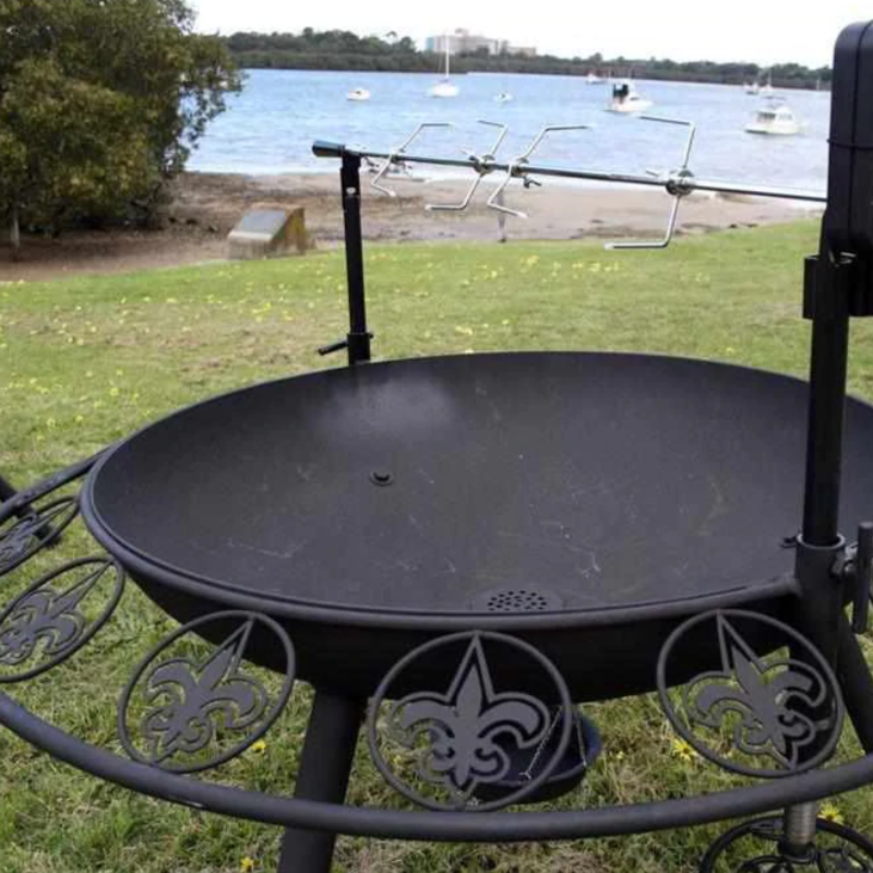 The Ultimate BBQ Fire Pit with a spit rotisserie and the Fleur De Lis pattern, on a lawn area beside a body of water with boats and trees in the background.