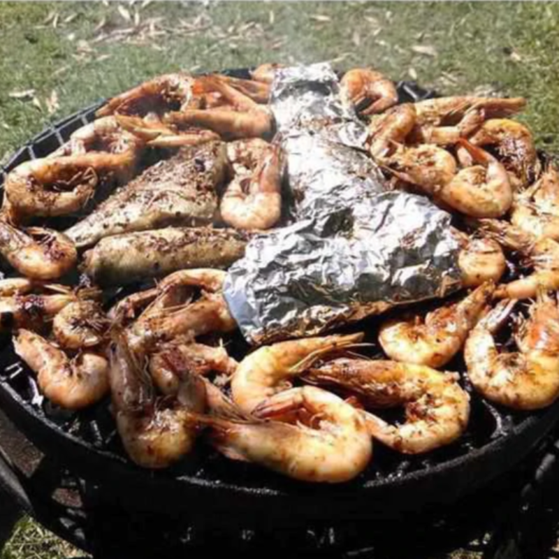 The Ultimate BBQ Fire Pit on a lawn with prawns being grilled in the shell, some fish and something else wrapped in foil also cooking.