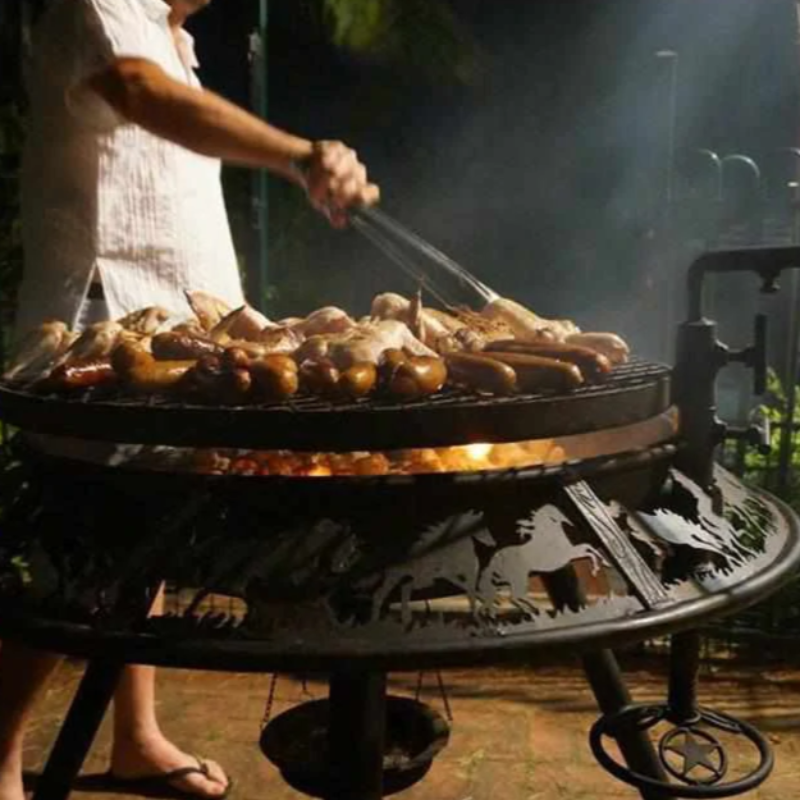 Sausages and chicken wings being cooked on the Stockman designed Ultimate BBQ Fire Pit, a man is turning the meat with BBQ tongs, on a paved area.
