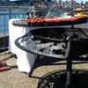 The stockman pattern Ultimate BBQ Fire Pit cooking up a feast on an outdoor cemented area besides a body of water and a bridge with buildings in the background.
