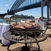 The stockman pattern Ultimate BBQ Fire Pit with meat cooking next to a table with food warmers set up in a cemented area besides a body of water and a bridge with buildings in the background.