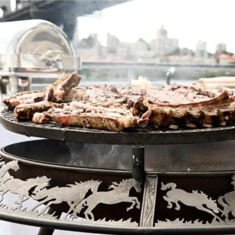 Close up view of the Ultimate BBQ Fire Pit with meat being grilled on the stockman design, buildings can be seen in the background.