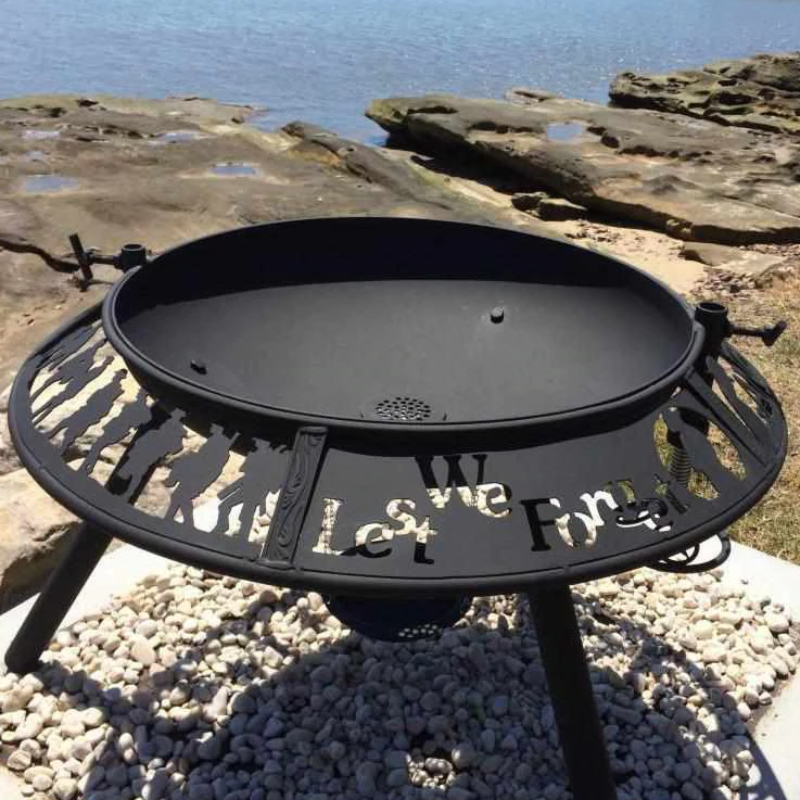 The Anzac patterned Ultimate BBQ Fire Pit with the words 'Lest We Forget' and soldiers cut out, set up on a gravel and rocky surround besides a lake.