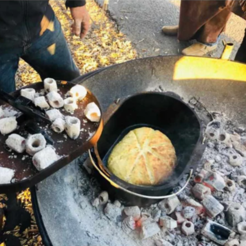 Baking damper in a tin inside the Ultimate BBQ Fire Pit, someone is also taking hot coals out of the fire pit with a metal scoop.